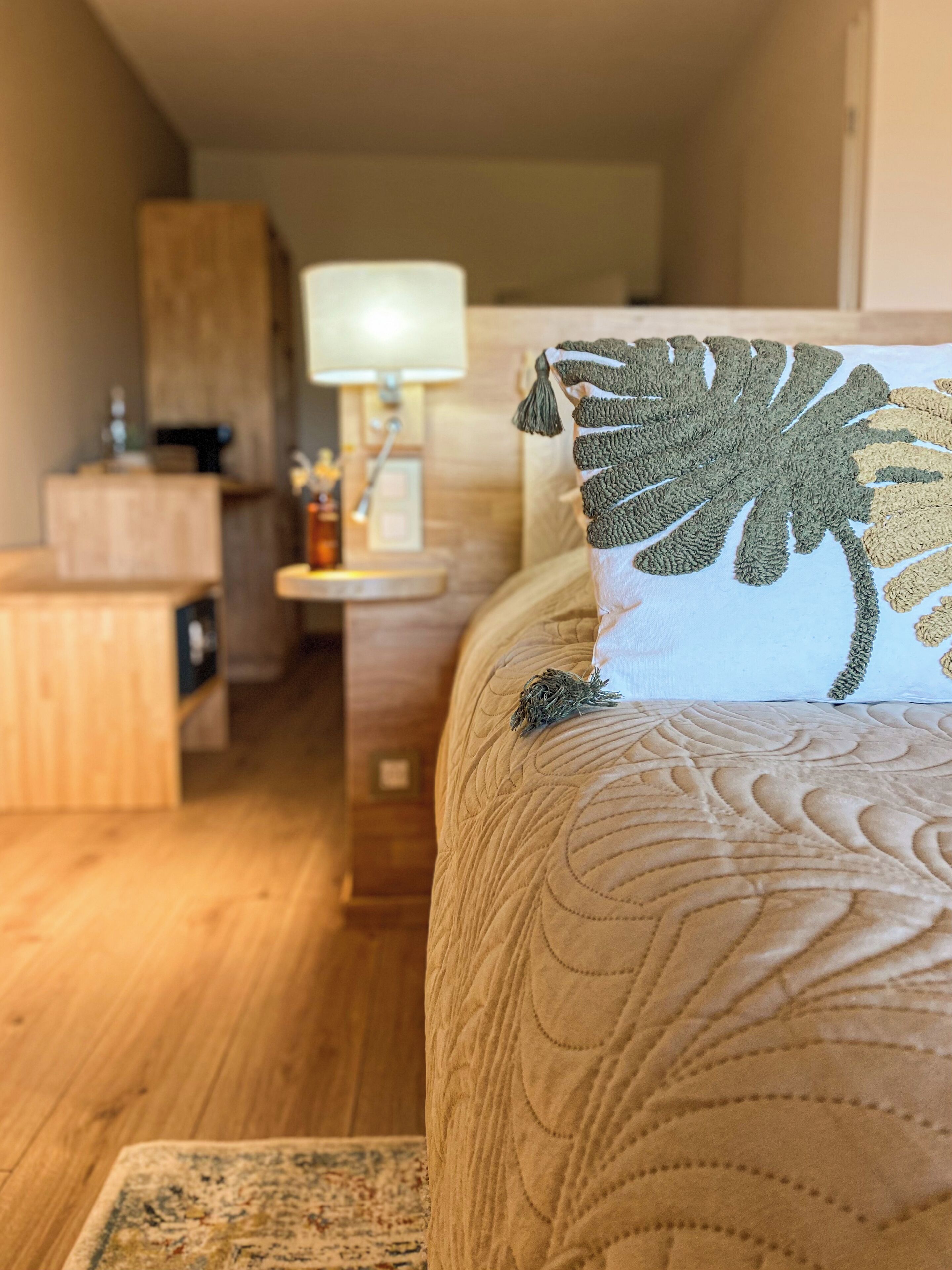 A neatly arranged hotel room with a bed, a bedside table, a lamp, and a small kitchenette in the background.