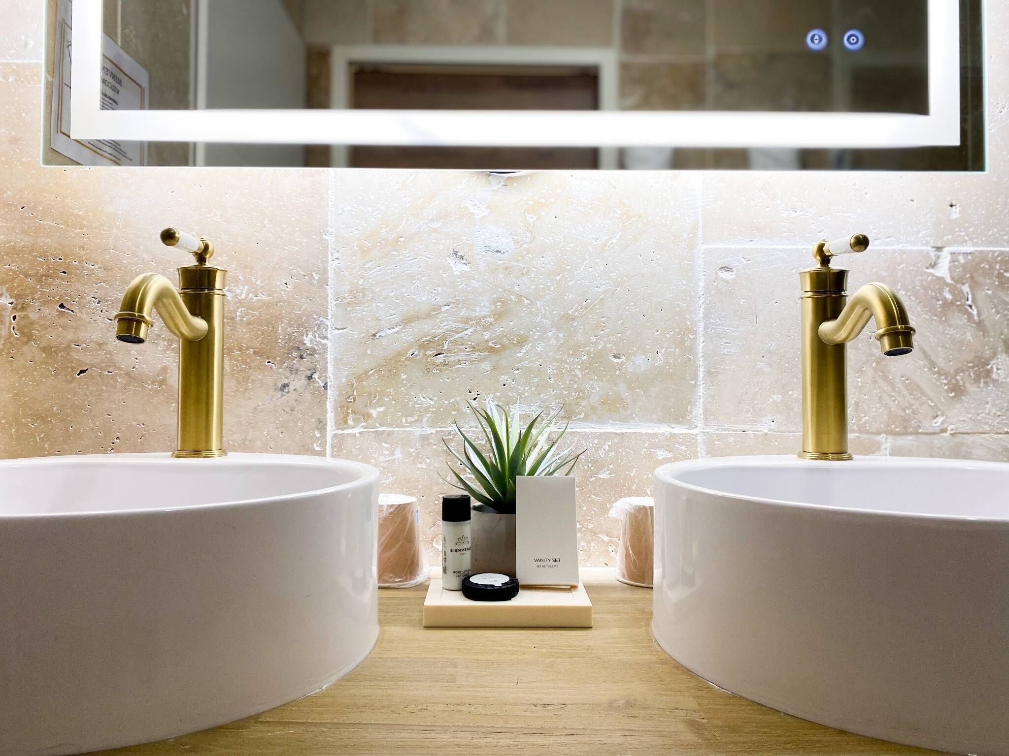 Un baño con dos grifos dorados, lavamanos blancos y un espejo arriba.