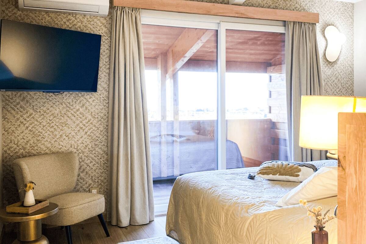 Habitación de hotel con cama, televisor montado en la pared, una silla, una mesita y ventana con cortinas.