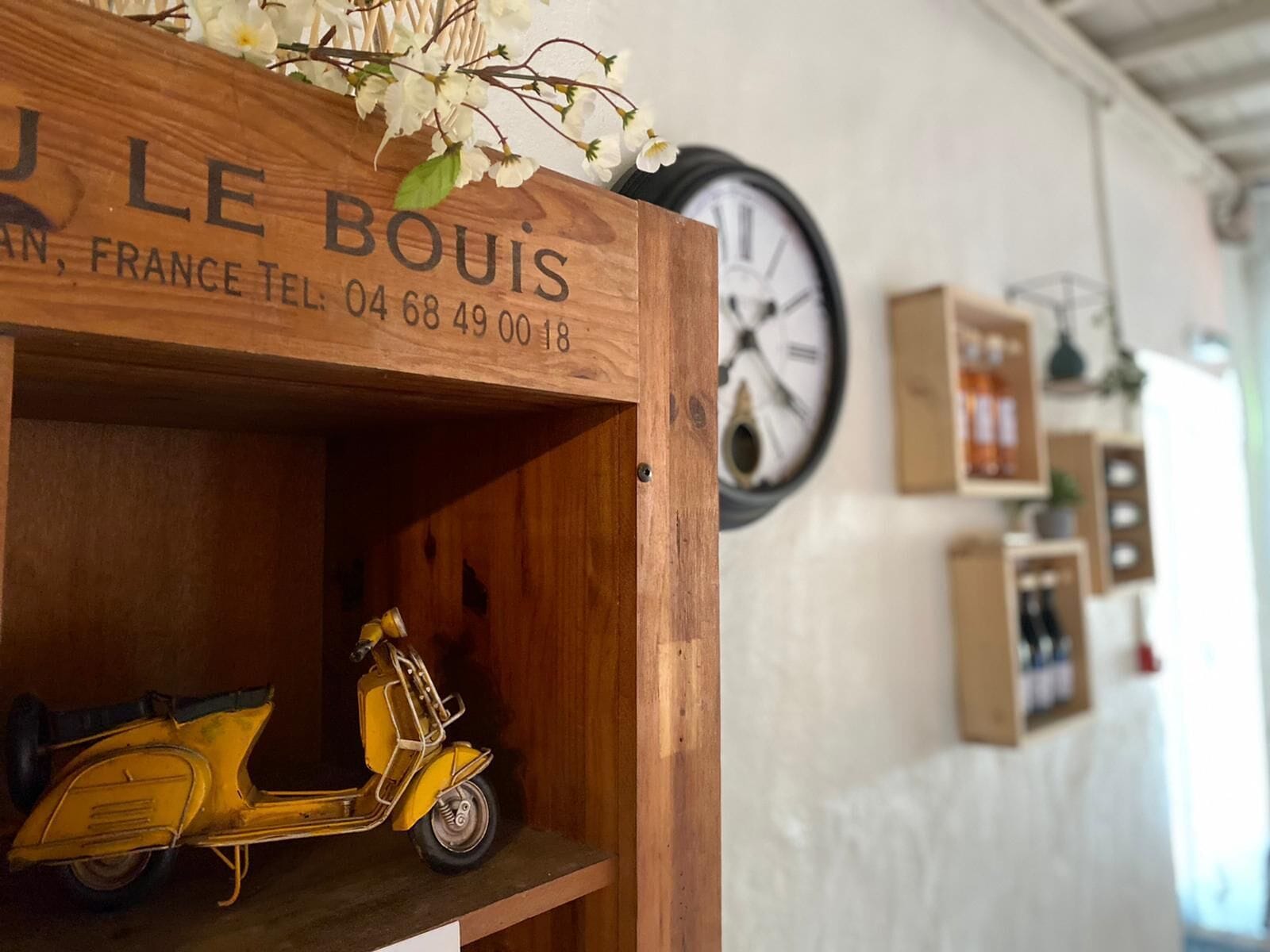 Une étagère en bois sur laquelle sont posés un scooter jaune d’époque et une enseigne indiquant : « LE BOUIS, FRANCE, TÉL. : 04 68 49 00 18 ».
