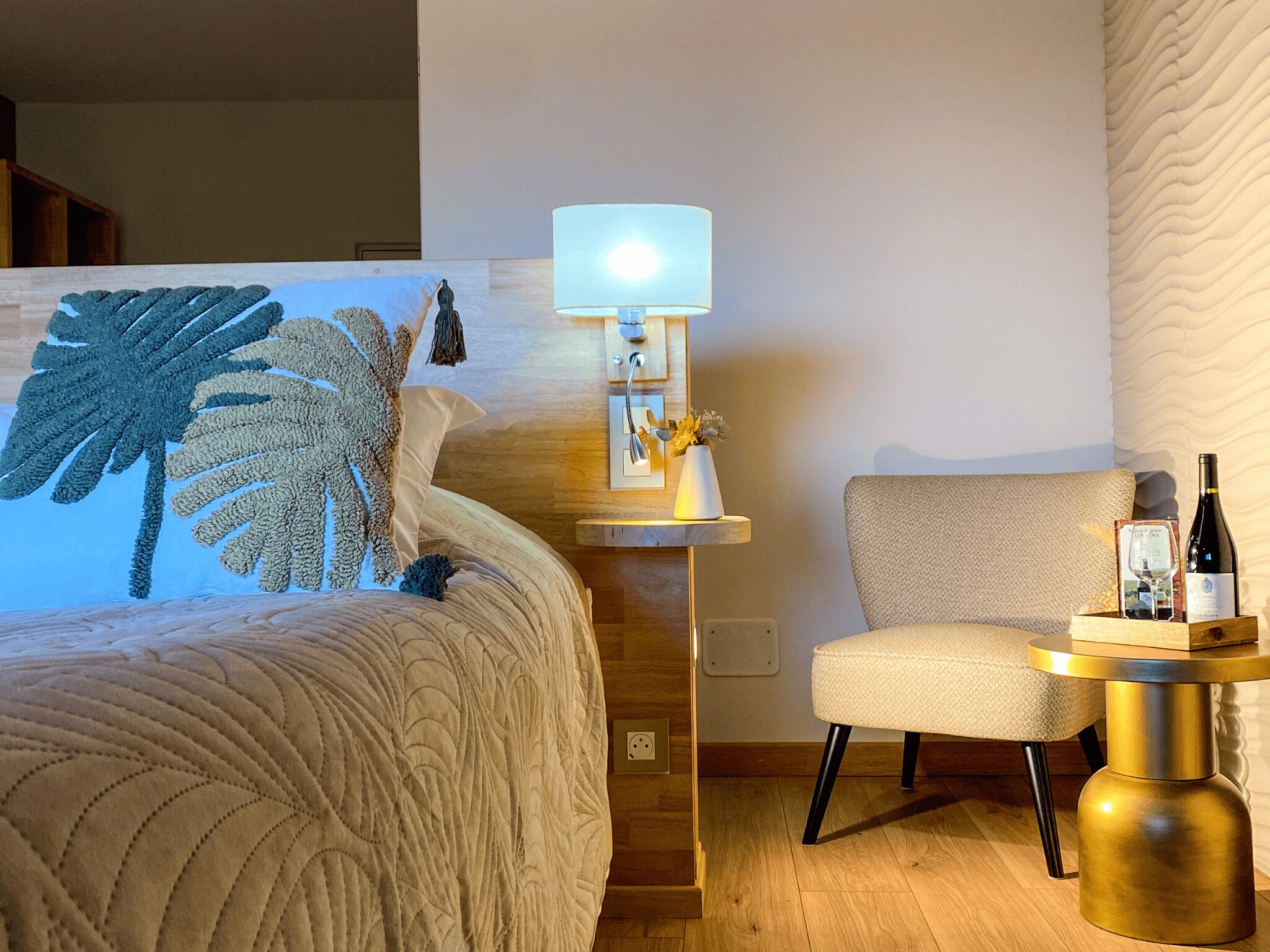 A hotel room with a bed, a chair, a lamp, and a small table with a bottle and glass.