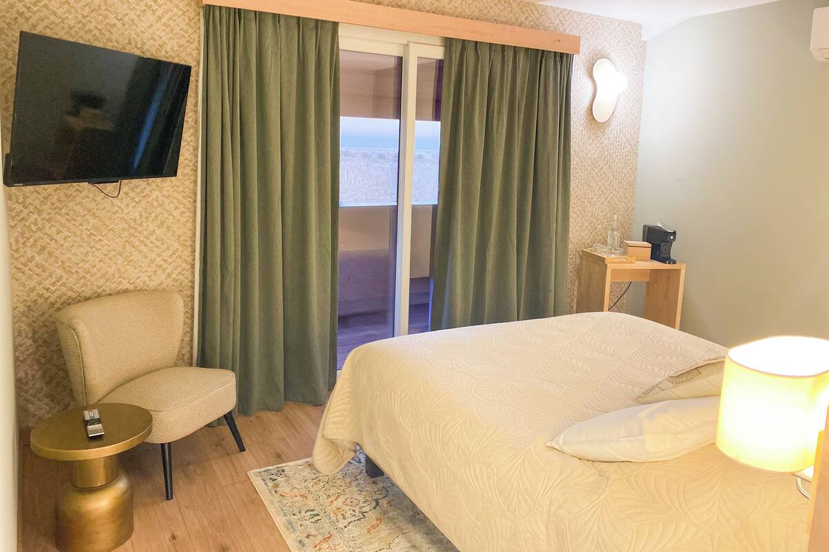 Habitación de hotel con cama, una silla, televisor, mesita y ventana con cortinas.