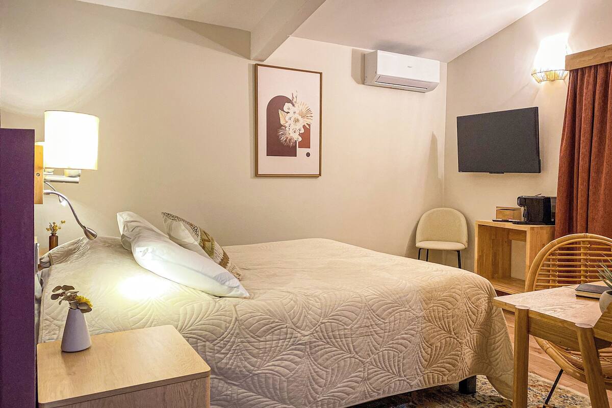 Habitación de hotel con cama, mesita de noche, lámpara, aire acondicionado, cuadro enmarcado, televisor y una silla.