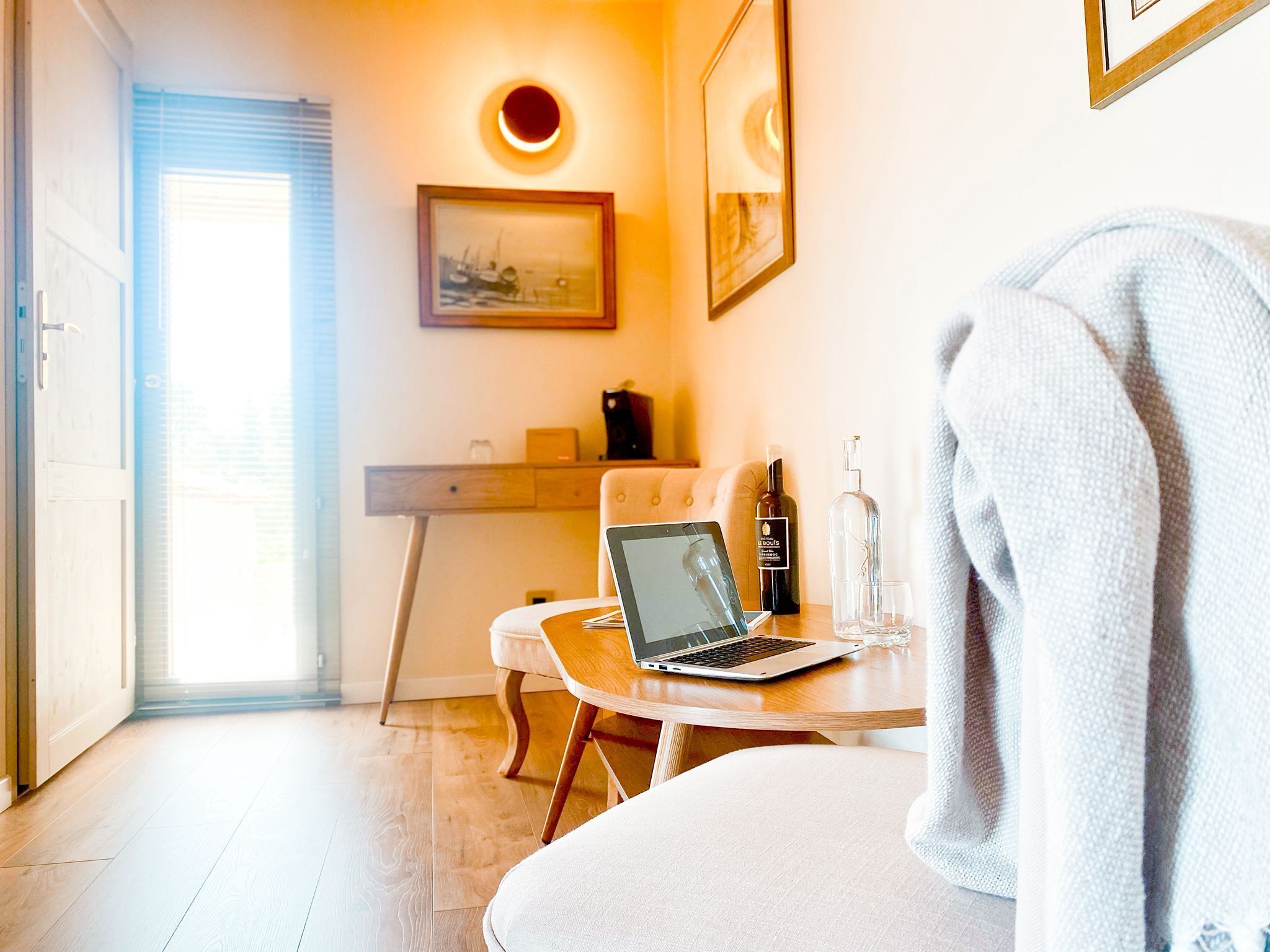 A cozy room with a laptop on a wooden table, a bottle of wine, and a glass. There is a chair and a bed with a blanket.