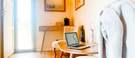 A cozy room with a laptop on a wooden table, a bottle of wine, and a glass. There is a chair and a bed with a blanket.