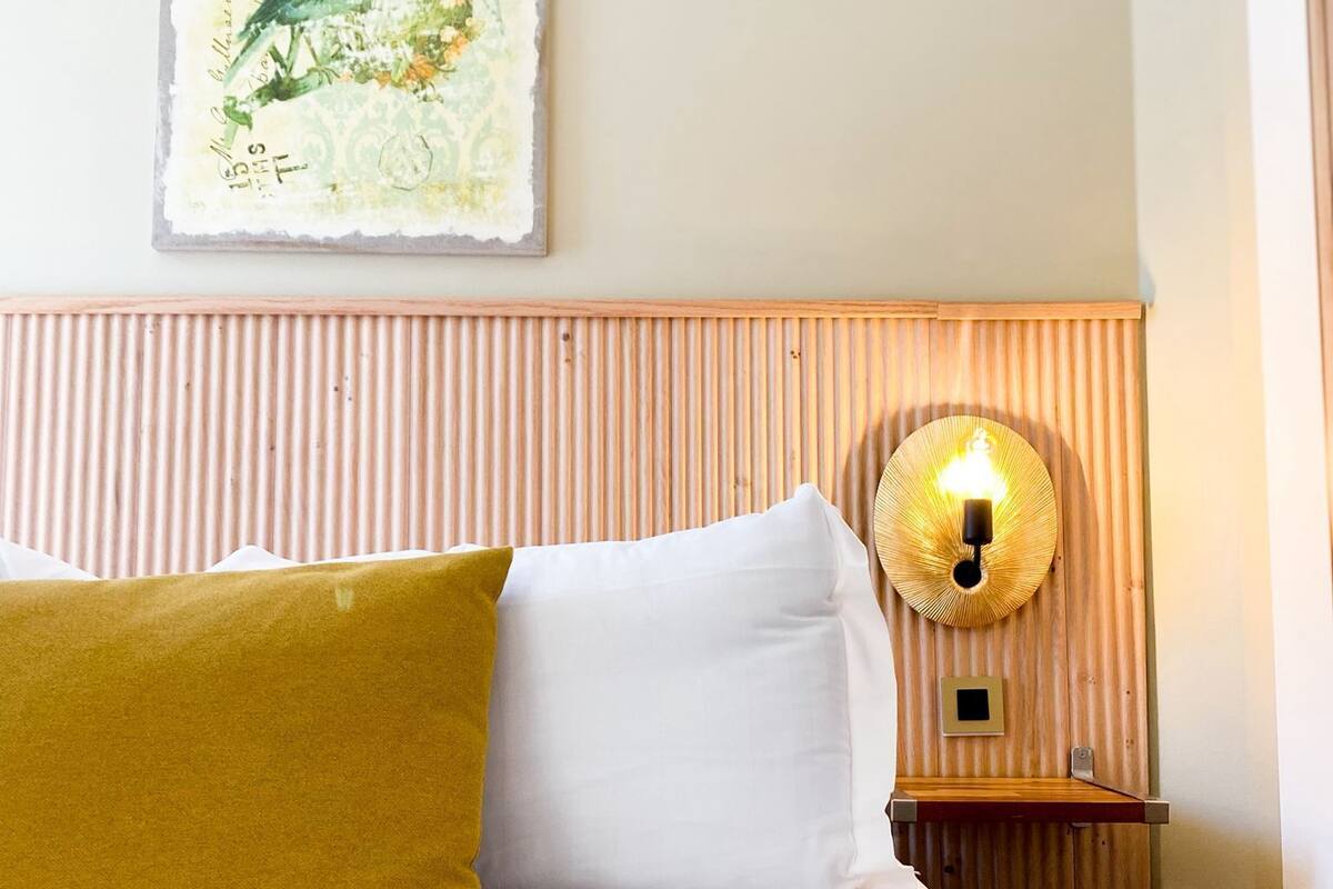 Una cama bien hecha con un libro abierto, una lámpara de pared y una obra de arte enmarcada sobre la cama.