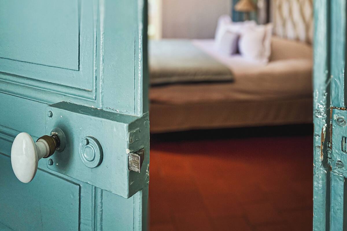 Una puerta verde azulado con un pomo blanco, que da a un dormitorio con una cama, almohadas y una mesita de noche.