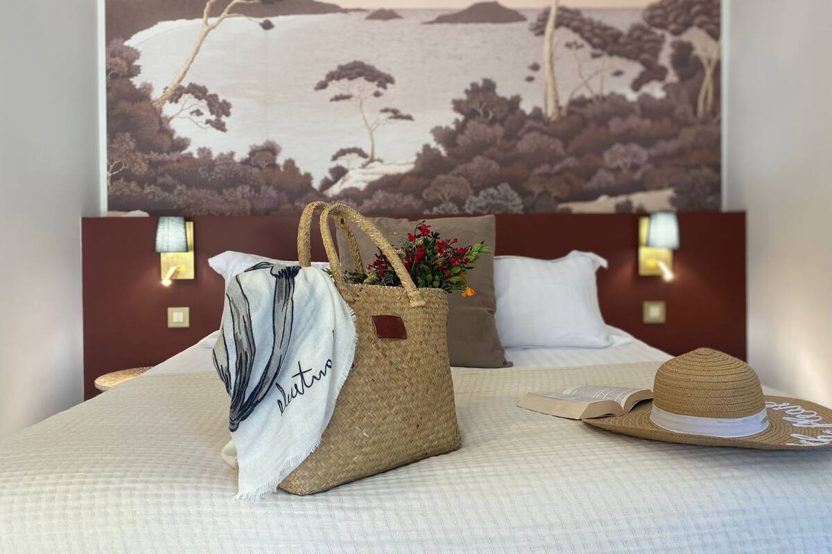 Una cama con una colcha blanca, un bolso de paja con flores y un sombrero de paja encima.
