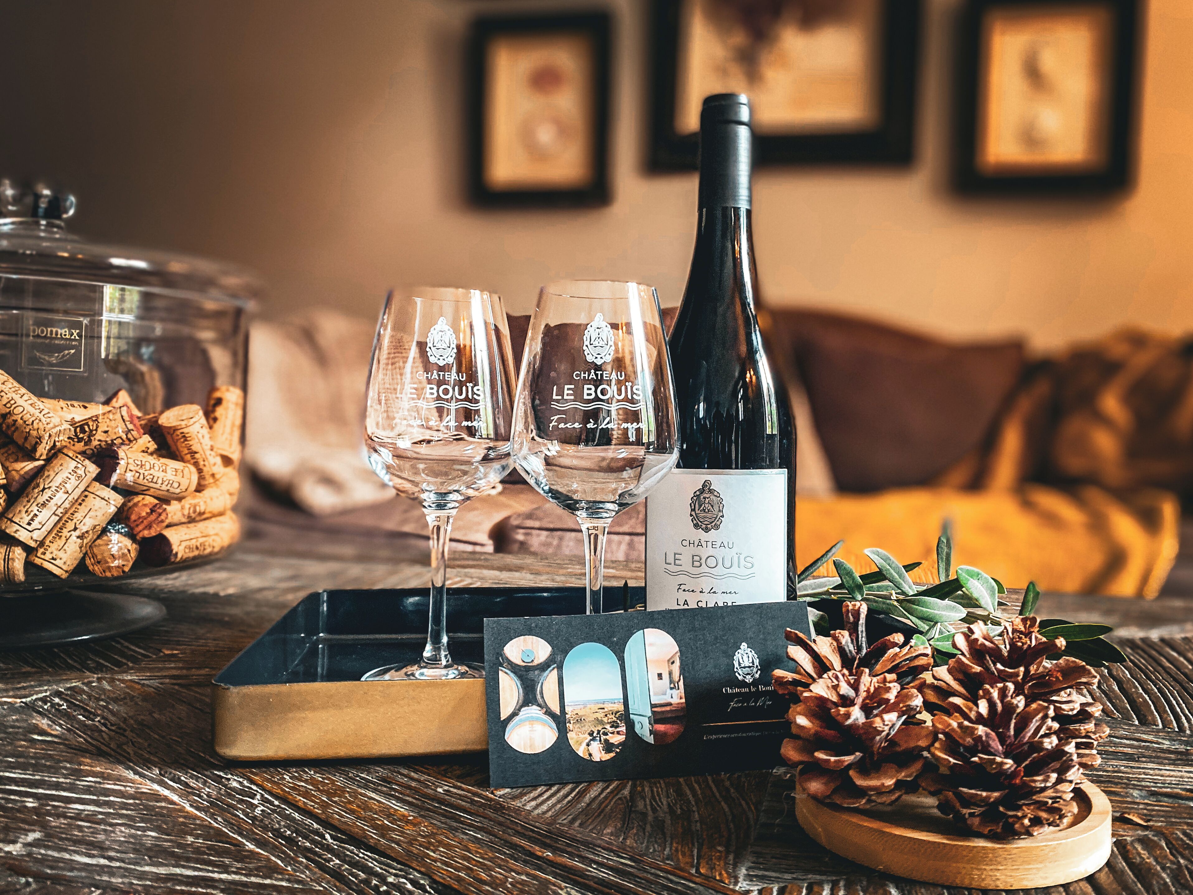 A bottle of Chateau Le Bouti, two wine glasses, and a box of wine corks on a wooden table.