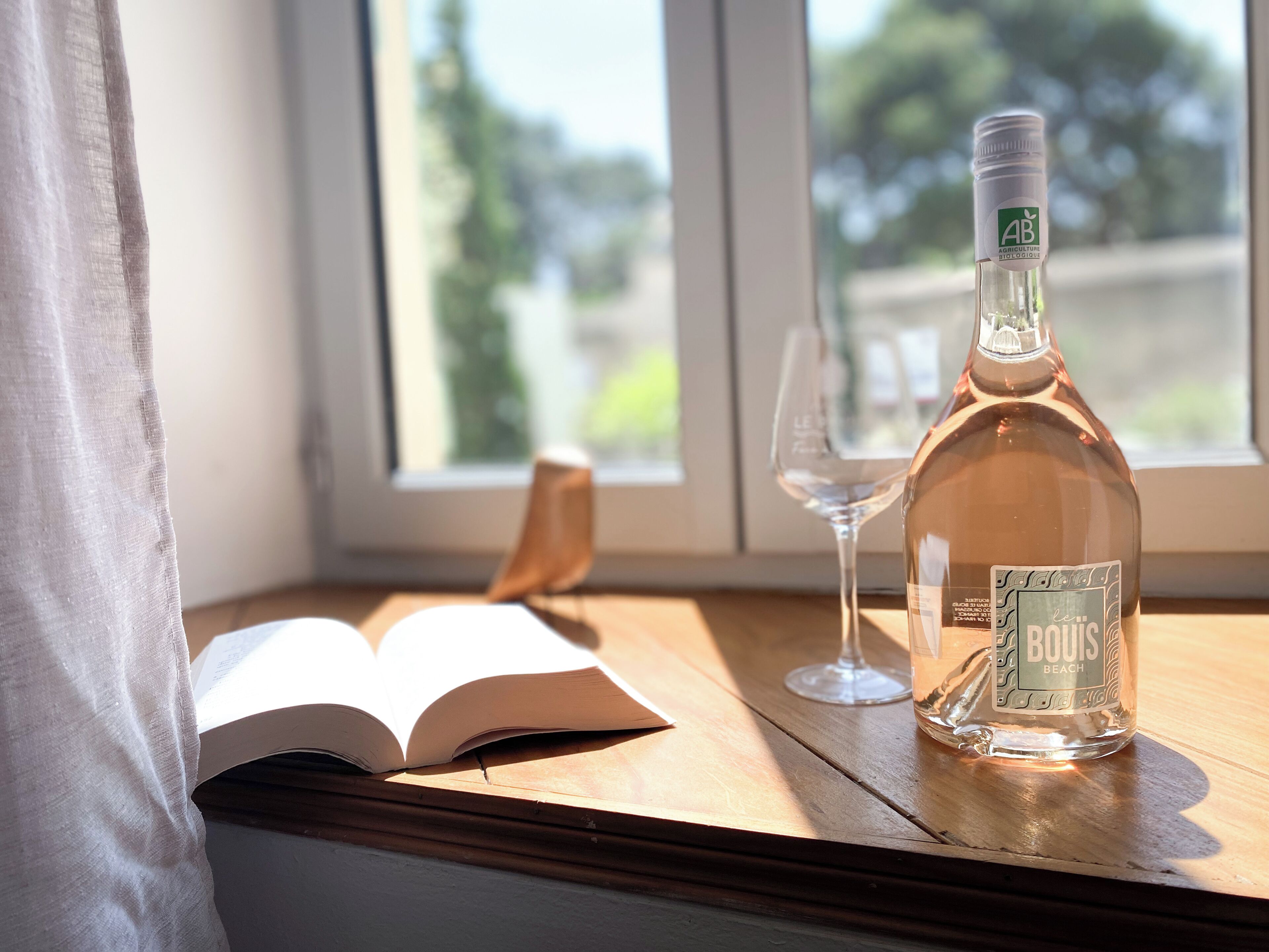 Una botella de alcohol y un libro abierto sobre una mesa de madera junto a una ventana.