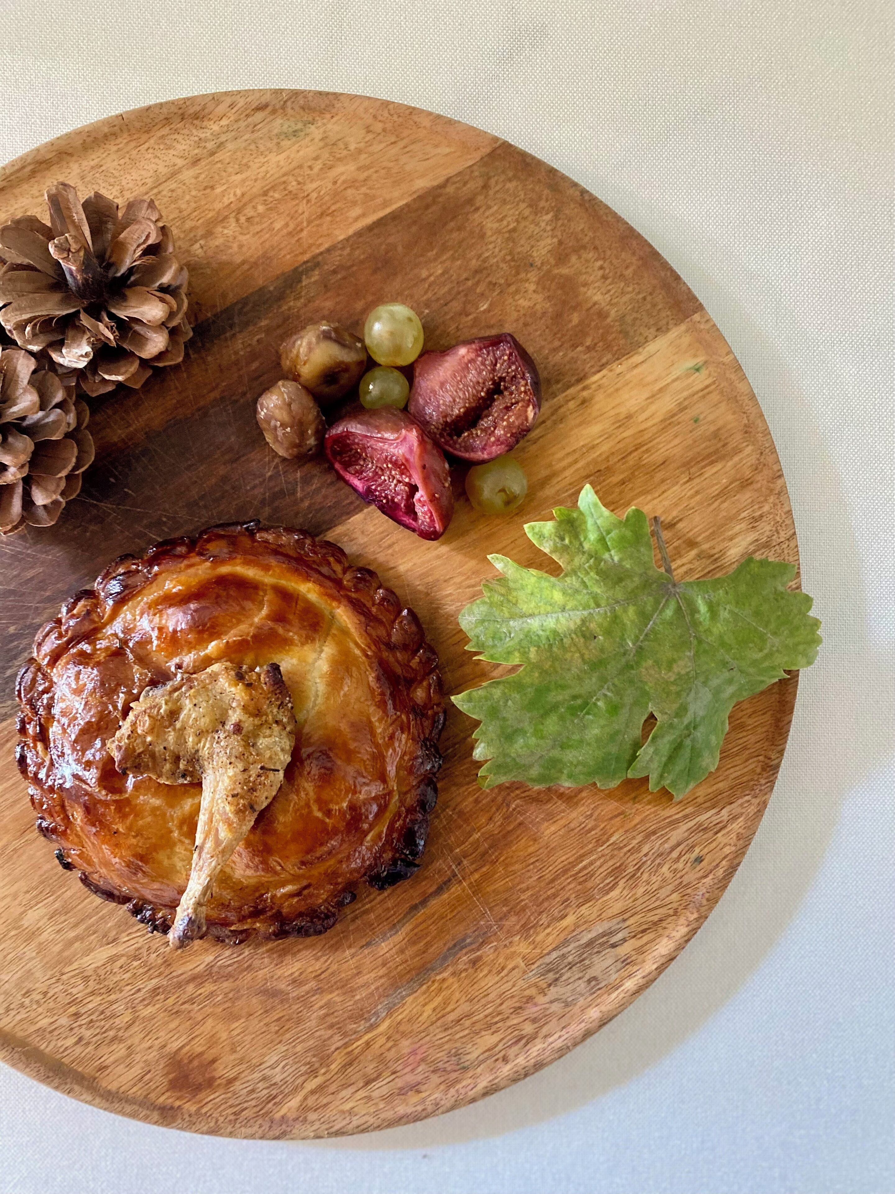 A wooden plate with a glazed meat dish, grapes, figs, and a pine cone.