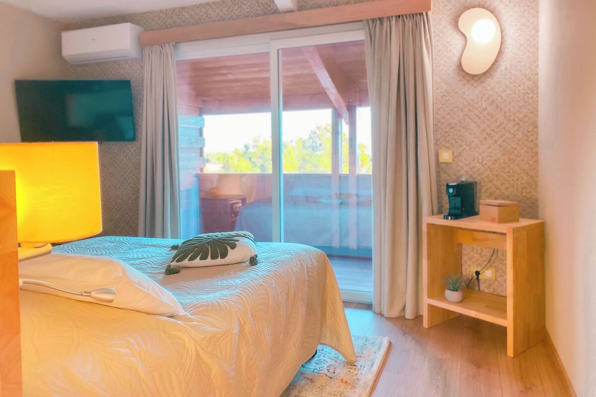 Un dormitorio con cama, una lámpara de noche, un pequeño estante de madera con una planta y un ventanal corredero que da a un espacio exterior.