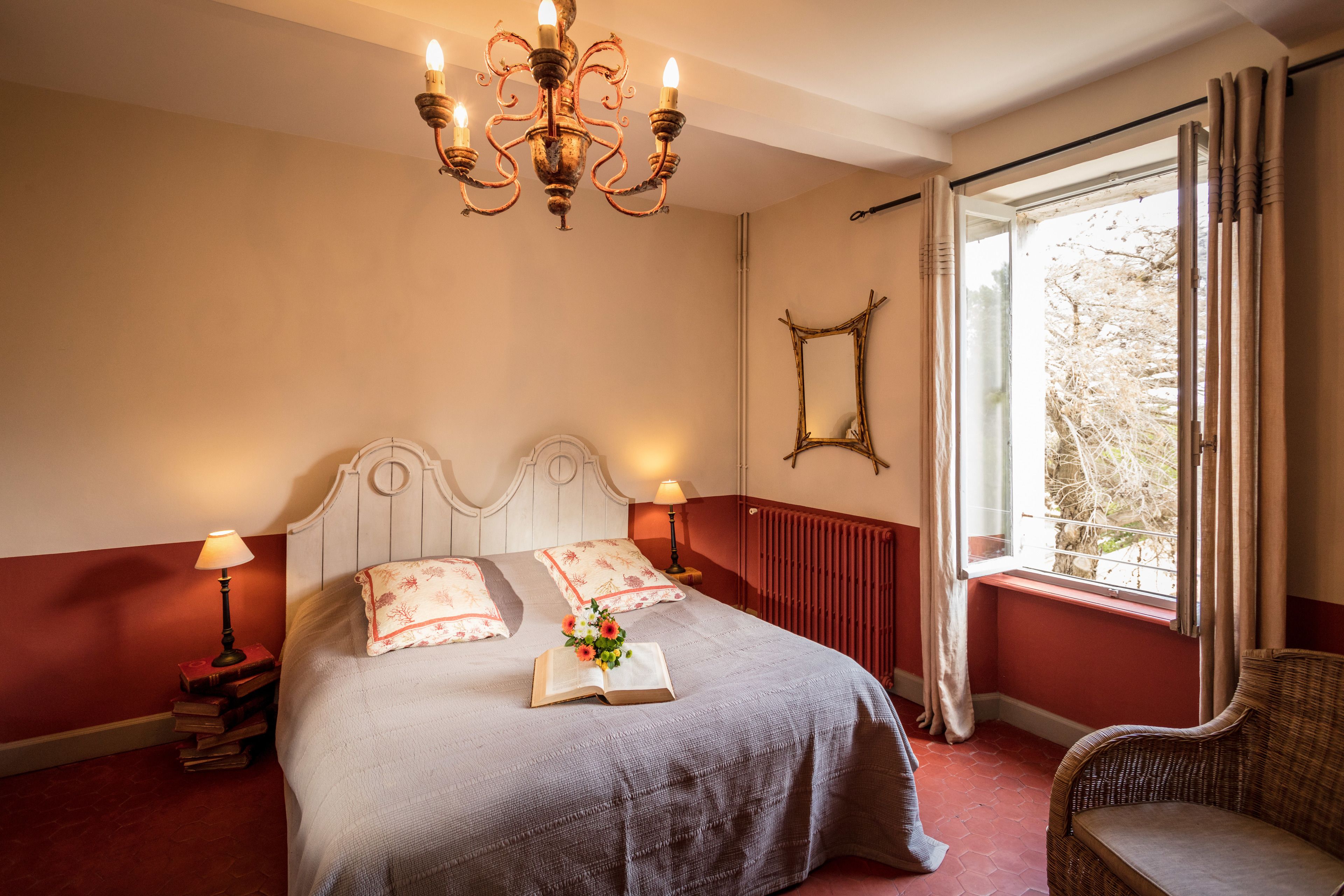 A neatly made bed with a book and flowers, a chandelier, a window with curtains, and a chair.