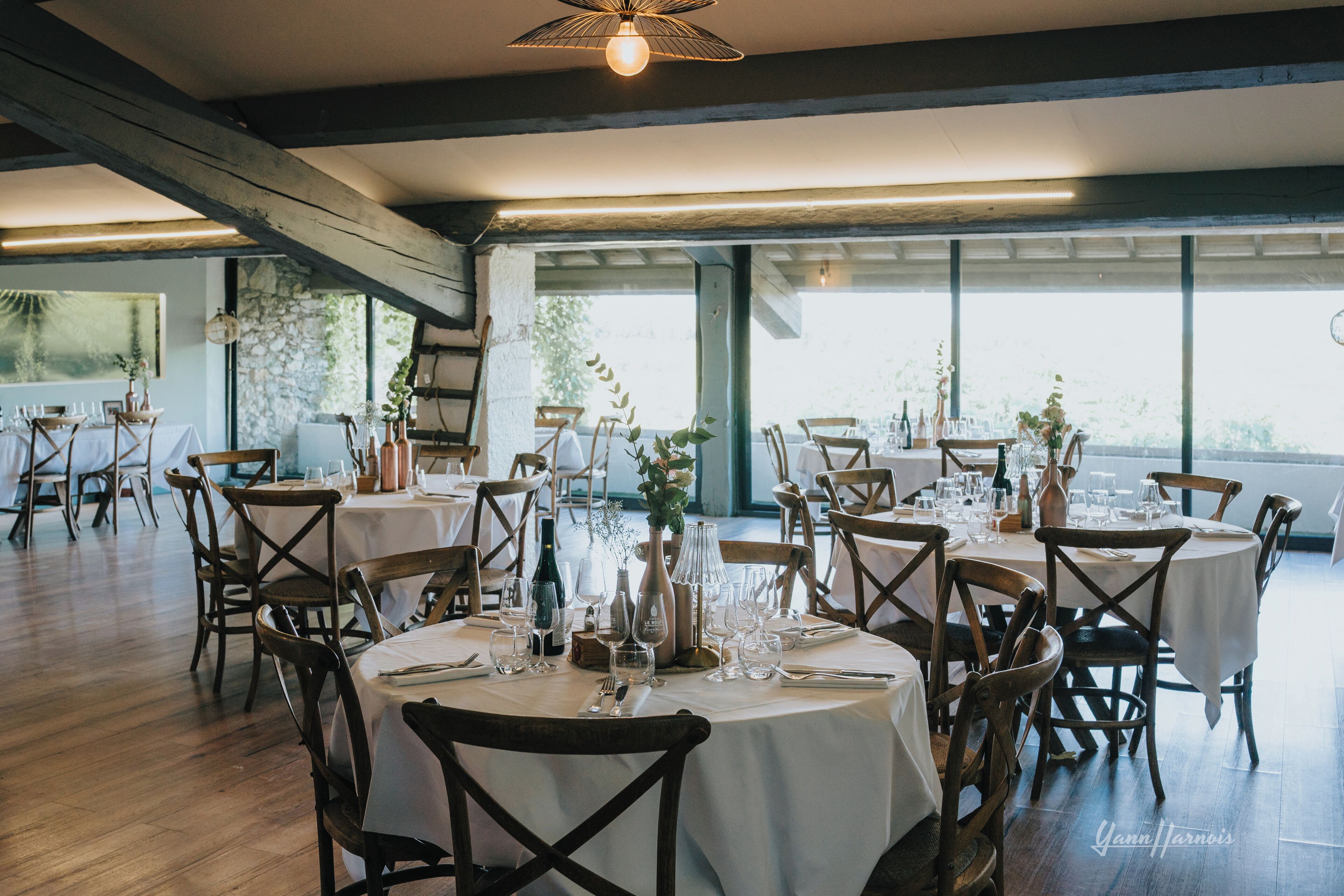A restaurant with round tables set for dining, wooden chairs, and a view through large windows.