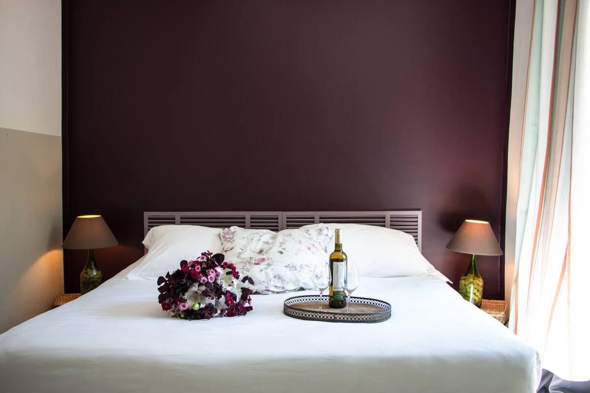 Una cama bien tendida con sábanas blancas, un cojín floral, una copa de vino y una bandeja con una botella.