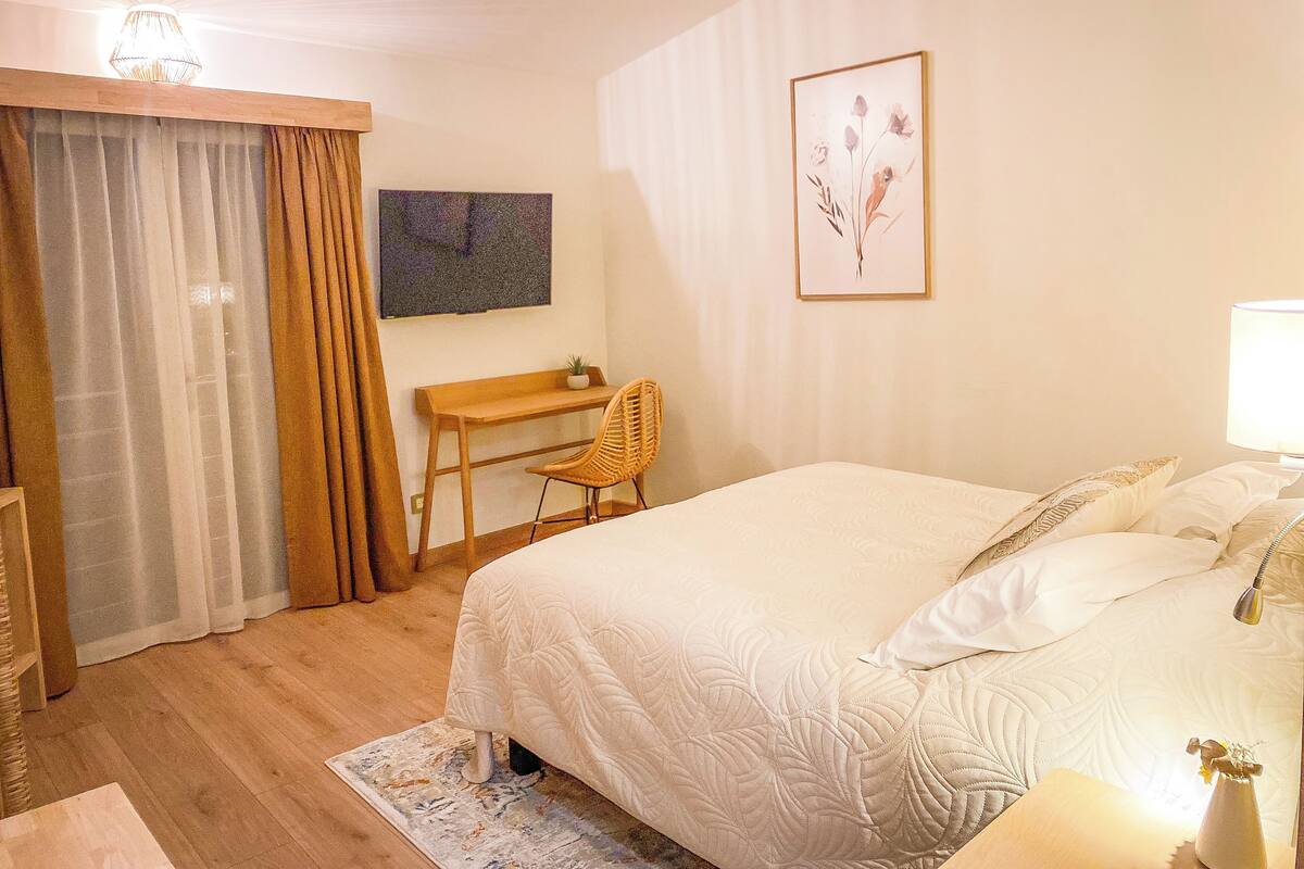Un dormitorio con cama, escritorio, silla, lámpara y un cuadro en la pared.