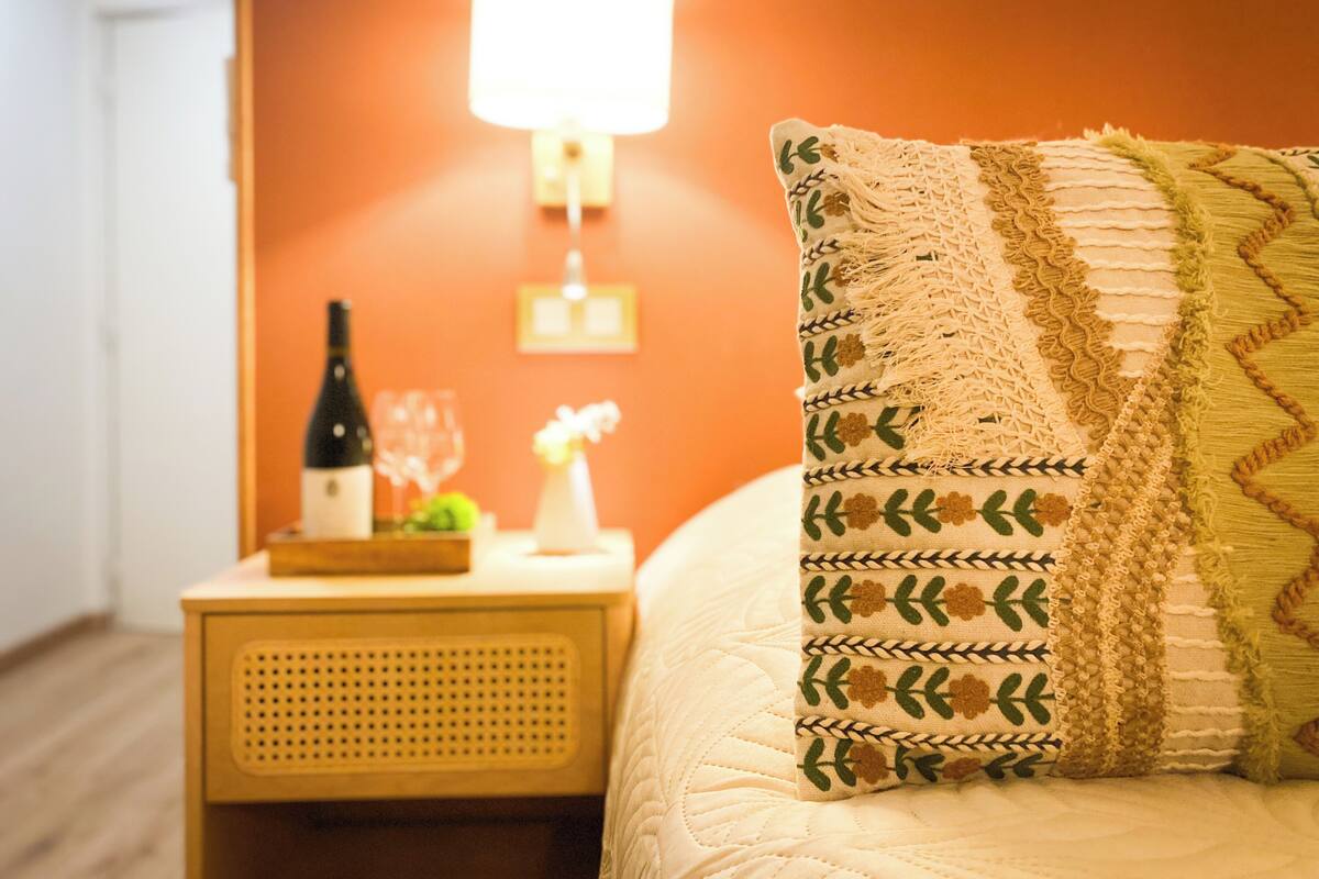 Una cama bien tendida con un cojín decorativo, una mesita de noche con una botella y un vaso, y una lámpara de pared.