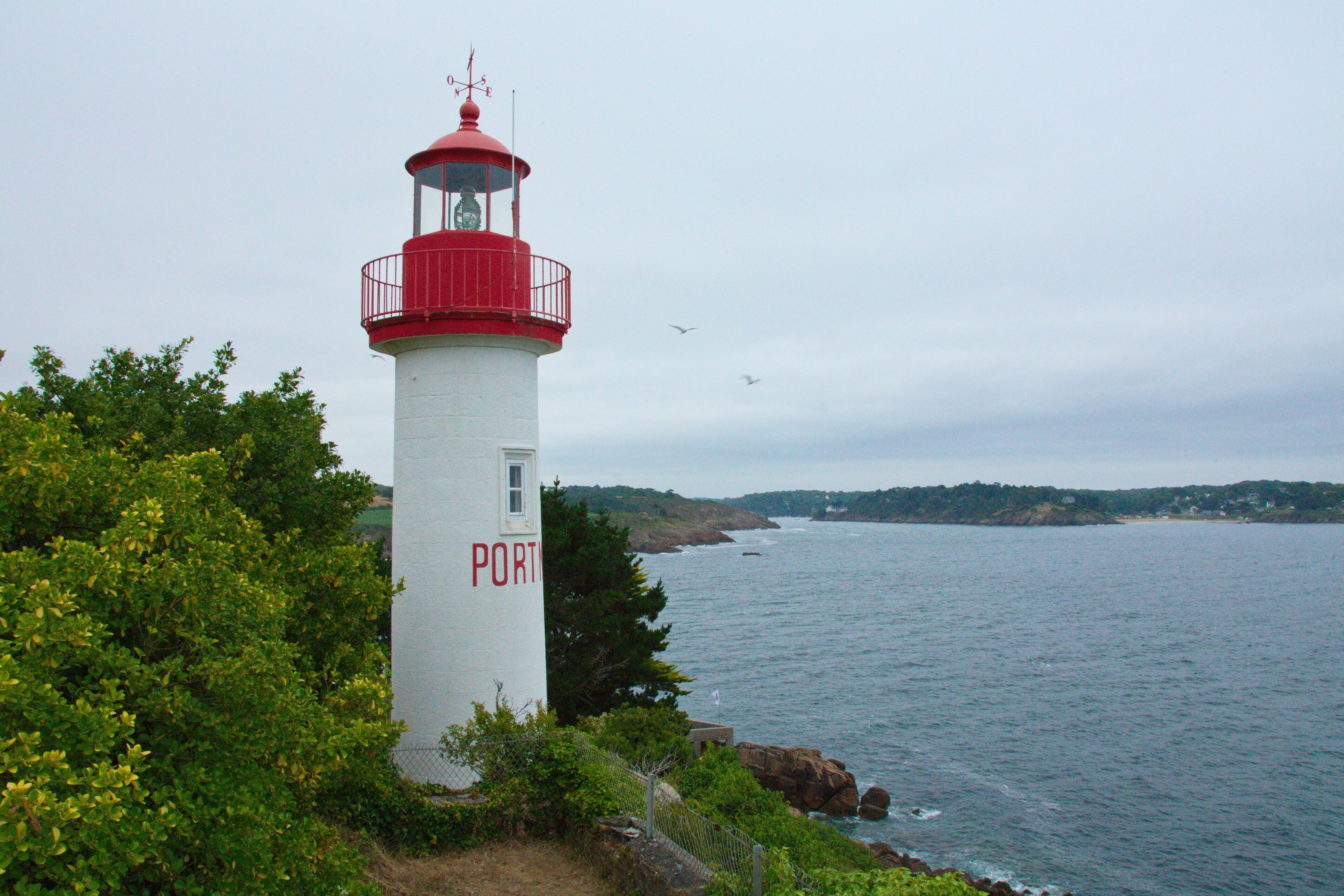 A lighthouse with a red top and white body, labeled "PORT", situated by a coastal area with greenery and a view of the sea.