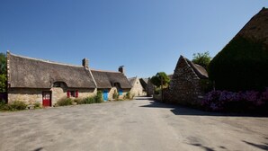 Une rue de campagne bordée de maisons en pierre, dont certaines sont coiffées de toits de chaume, sous un ciel d’un bleu éclatant.