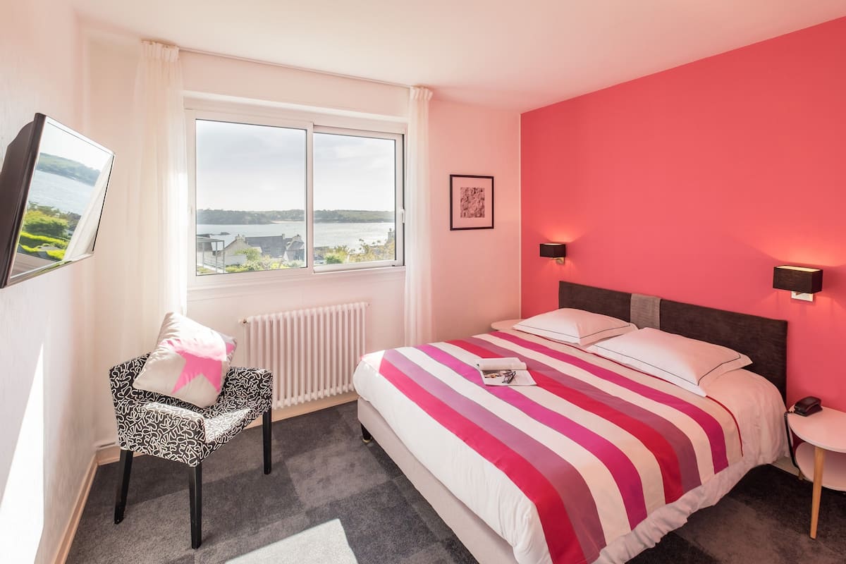 Une chambre d’hôtel avec un grand lit, une chaise, une vue sur l’eau et un tableau au mur.