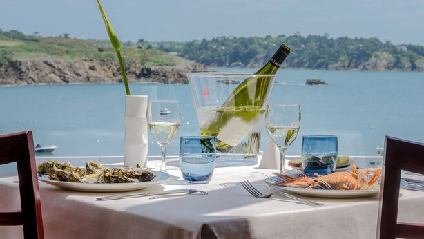 Une table dressée avec des assiettes de fruits de mer, des verres et une bouteille de vin, donnant sur une étendue d’eau.