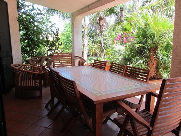 Une terrasse couverte avec une table et des chaises en bois, entourée d’une végétation luxuriante et d’un palmier.