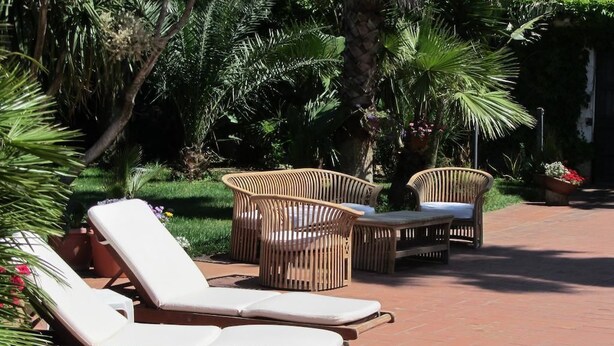Un patio con sillones de descanso y mobiliario de mimbre, rodeado de abundante vegetación y un camino de ladrillo.