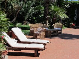 Un patio con sillones de descanso y mobiliario de mimbre, rodeado de abundante vegetación y un camino de ladrillo.