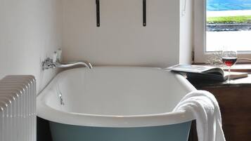 A bathroom with a freestanding bathtub, a radiator, a shelf with plants, and a window with a view of the sea.
