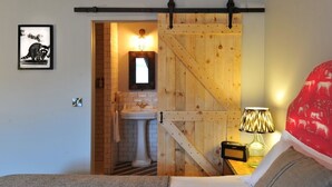 Un dormitorio con cama, mesita de noche con lámpara, puerta de madera y baño visible a través de una puerta abierta.