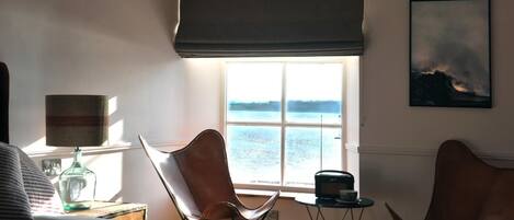 Una habitación con una cama, dos sillas, una mesa con una radio, una lámpara y un ventanal con vista al agua.