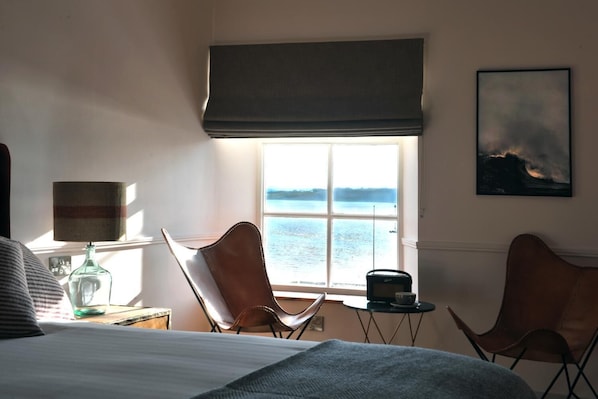 A room with a bed, two chairs, a table with a radio, a lamp, and a window with a view of water.