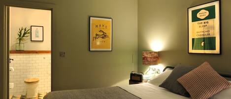 Un dormitorio con cama, lámpara de noche, un libro enmarcado en la pared y un baño visible a través de una puerta abierta.