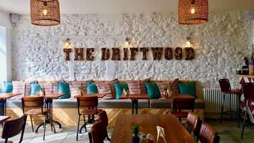 A cozy restaurant interior with a stone wall, wooden furniture, and a sign reading "THE DRIFTWOOD".
