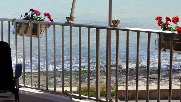 A balcony with metal railings overlooking a rocky shoreline with waves. There are flower pots with red flowers on the railing. - Sea View Apartment (Ragusa)