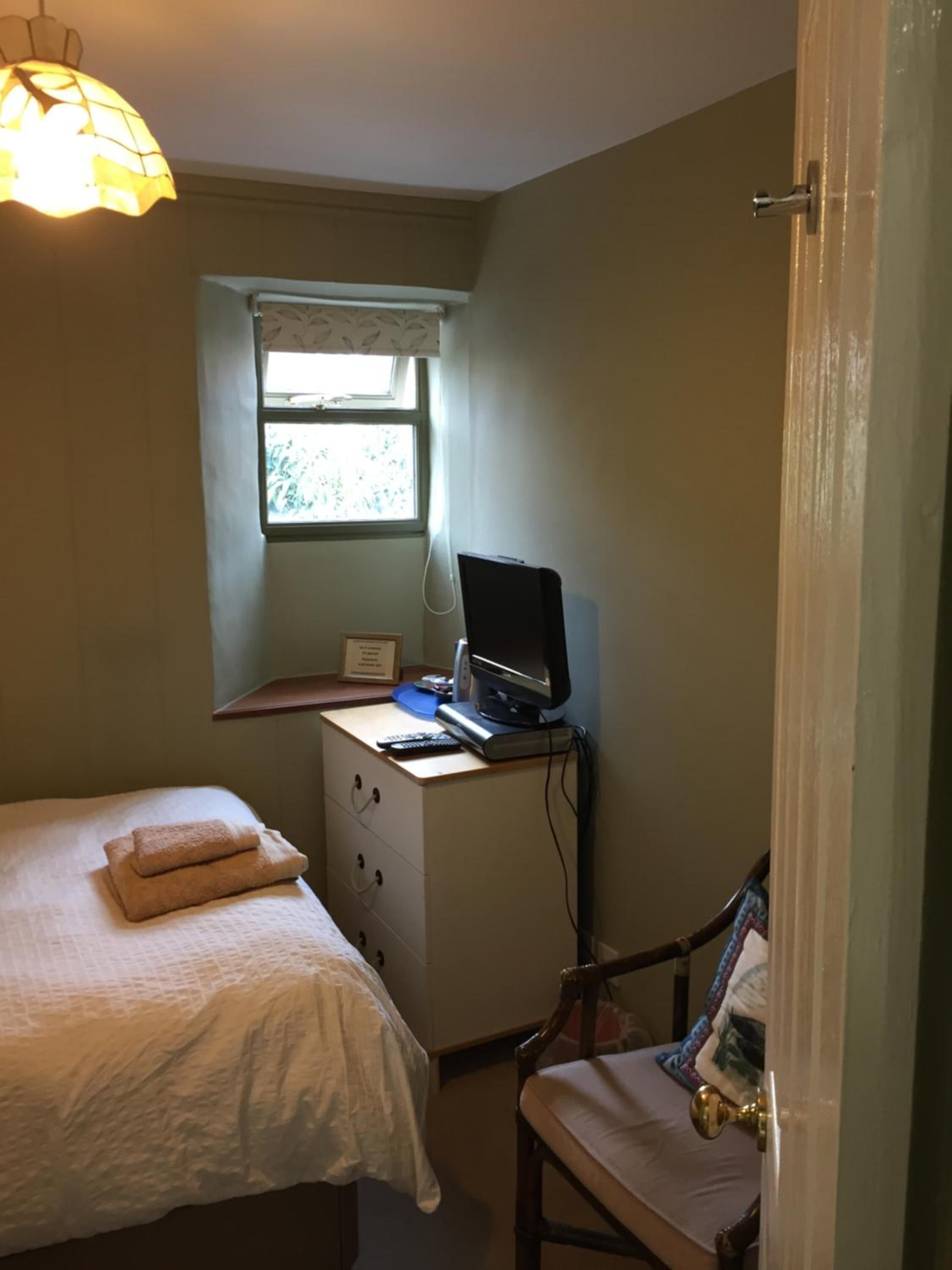 Ein Schlafzimmer mit einem Bett, einem Schreibtisch mit Computer, einem Fenster und einer Tür.