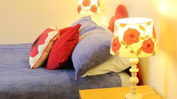 A bed with blue bedding, red and white pillows, and a white lamp with a floral lampshade on a wooden nightstand.