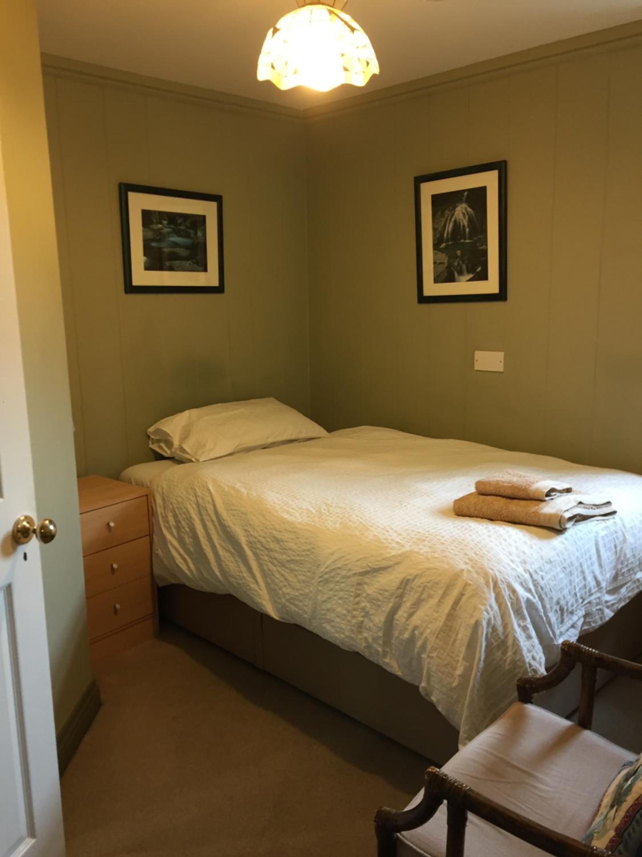 Een kleine, verzorgde slaapkamer met een enkel bed, een houten nachtkastje, een stoel en twee ingelijste foto's aan de muur.
