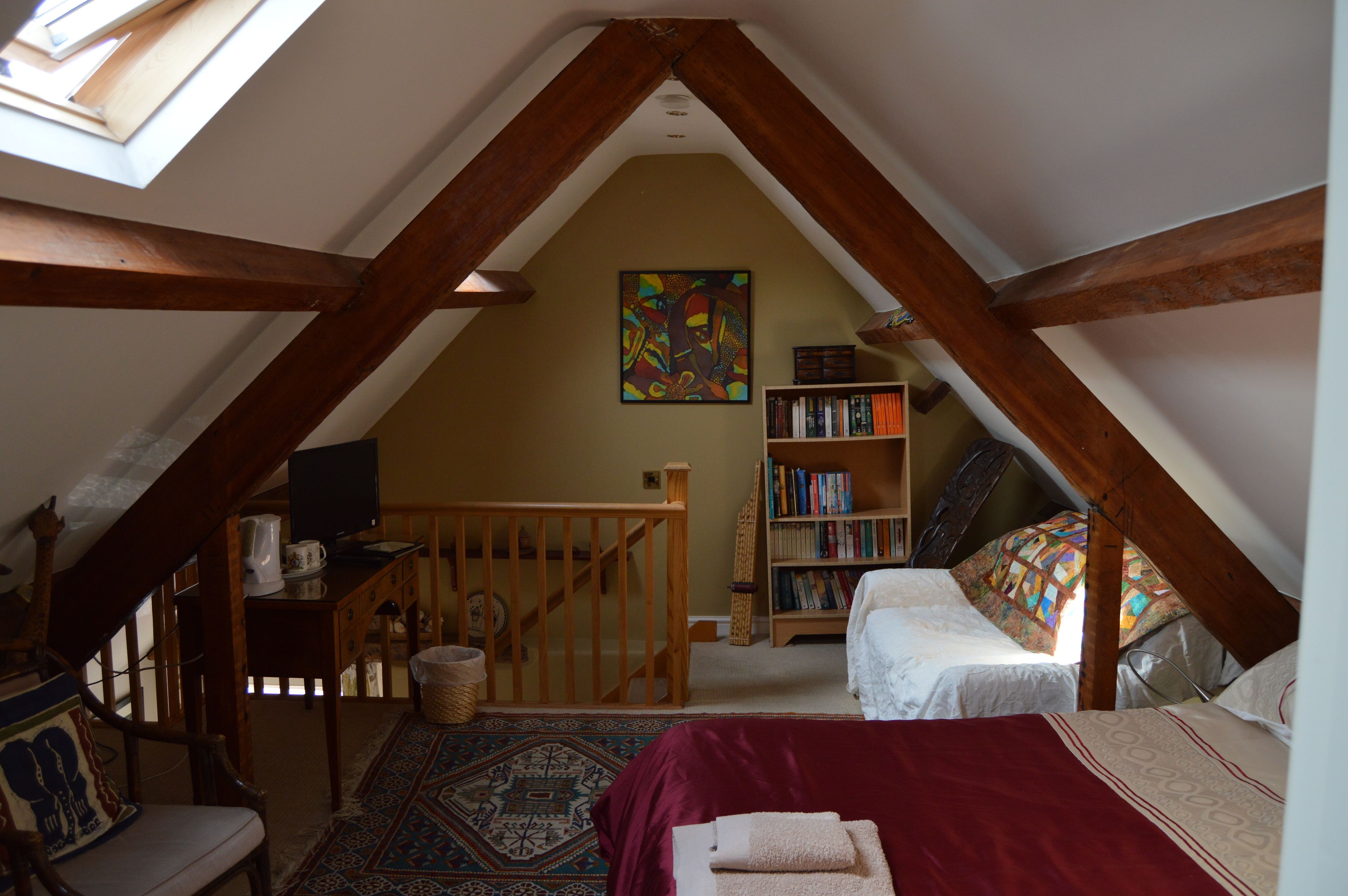A cozy attic bedroom with a sloped ceiling, a wooden staircase, a bookshelf, a bed with a red blanket, and a colorful abstract painting on the wall.