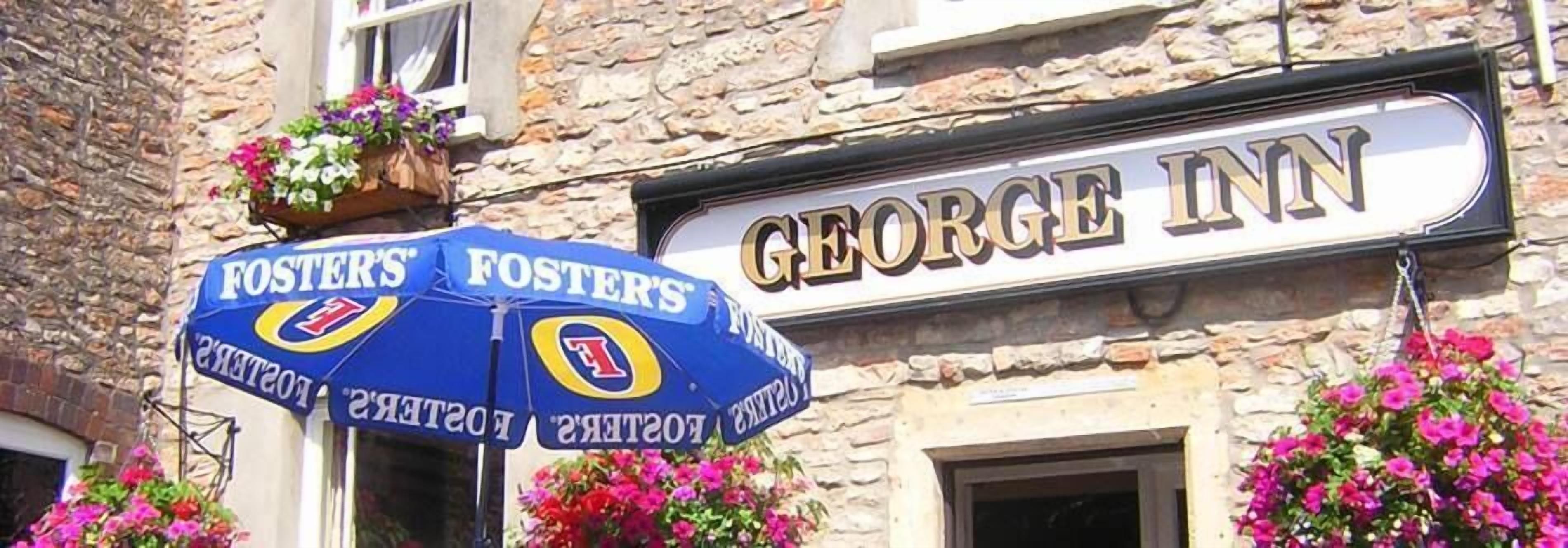 A building with a sign reading "GEORGE INN" and a blue umbrella with "FOSTER'S" branding.