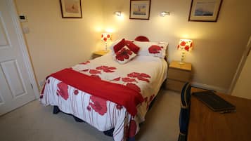 Una cama bien tendida con una colcha floral y almohadas rojas, dos lámparas de noche y una computadora portátil sobre una mesita de madera.