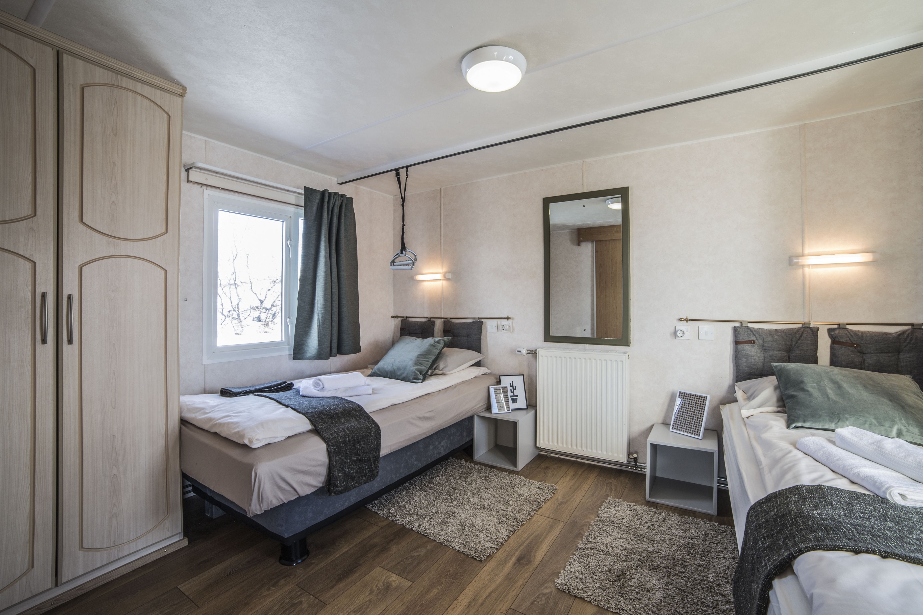 A bedroom with two beds, a wardrobe, a window with curtains, bedside tables, and a mirror.
