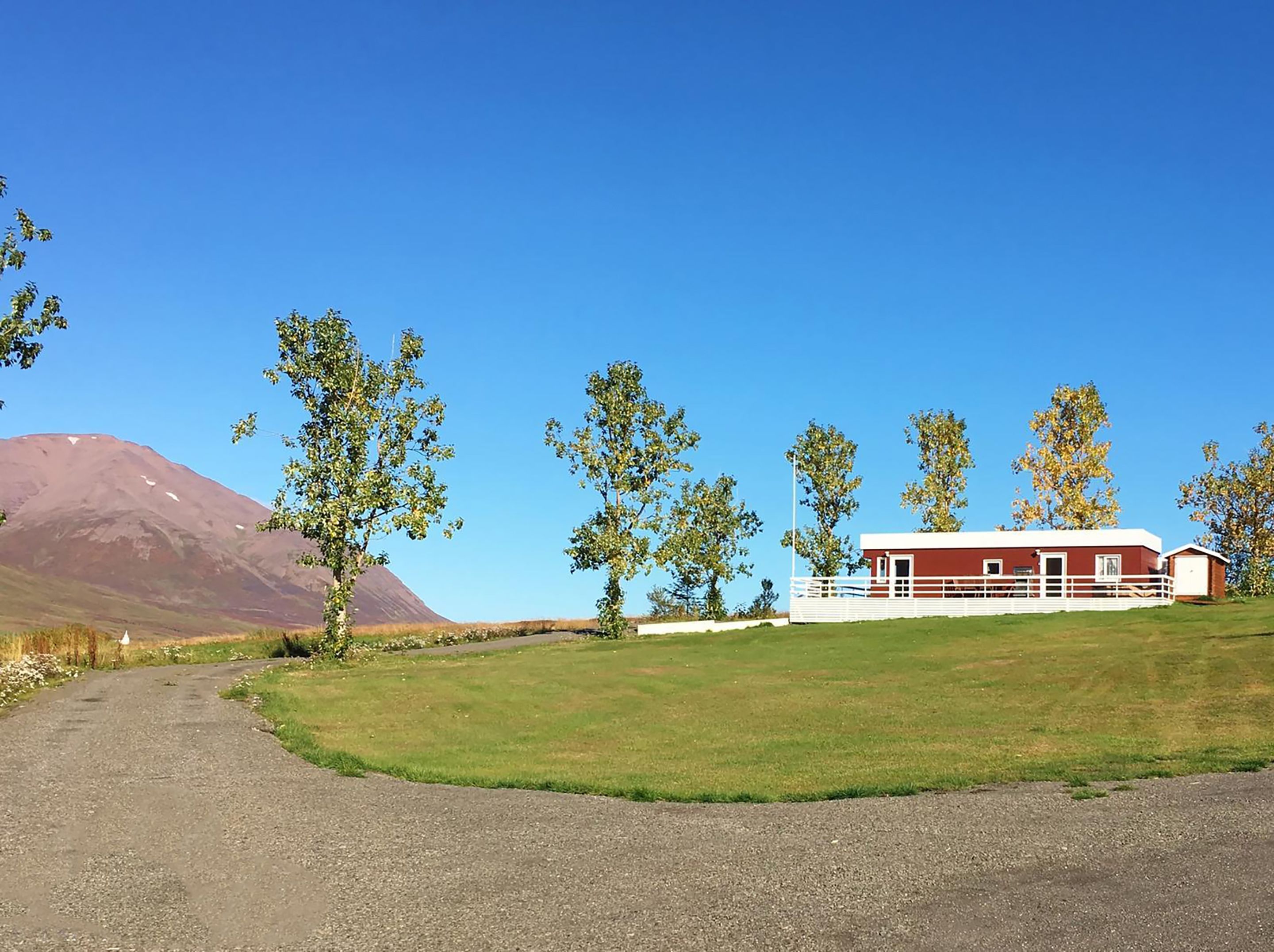 Un paysage rural avec un chemin de gravier menant à un bâtiment rouge, délimité par une clôture blanche, entouré d’arbres et avec une montagne en arrière-plan.