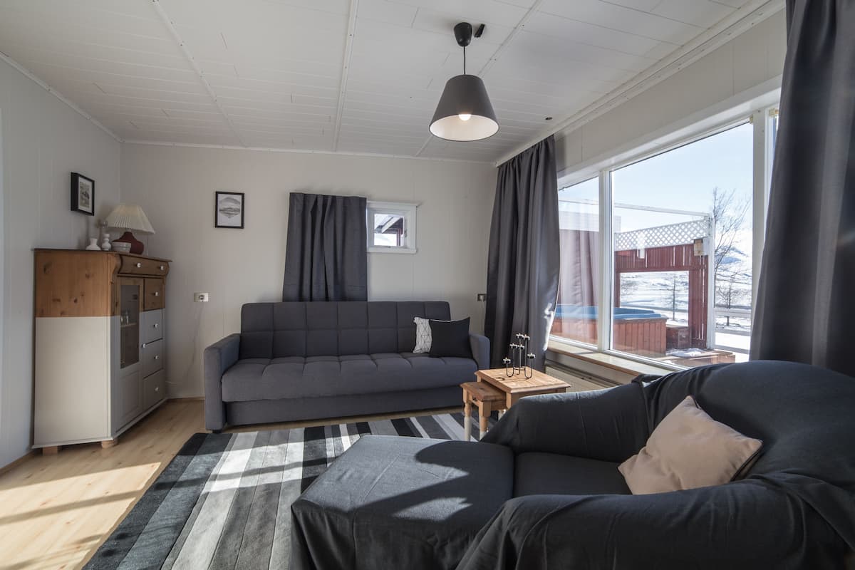 Ein modernes Wohnzimmer mit einem grauen Sofa, einem kleineren schwarzen Sofa, einem Holztisch und einem großen Fenster mit Vorhängen.