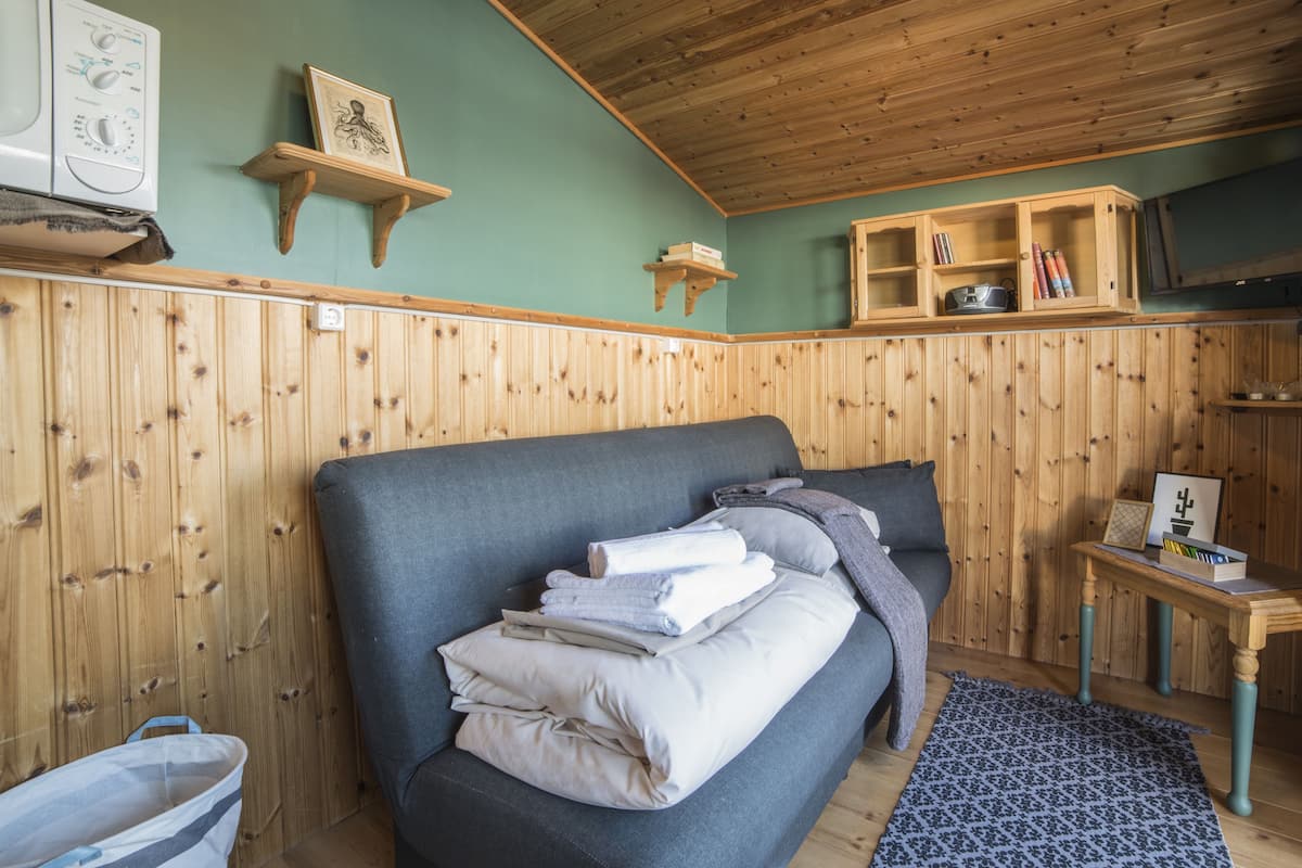 Ein gemütliches Zimmer mit einer holzverkleideten Wand, einem grauen Sofa mit Handtüchern, einem kleinen Holztisch mit Büchern und einem Bilderrahmen sowie einem blauen Teppich mit Muster.