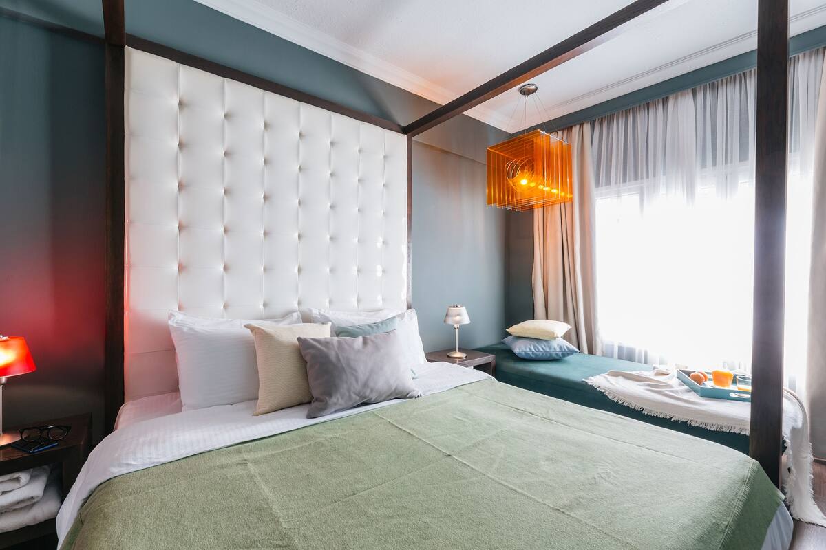 A hotel room with a large bed, a nightstand, a bedside table, a lamp, a window with curtains, and a view of the outdoors.