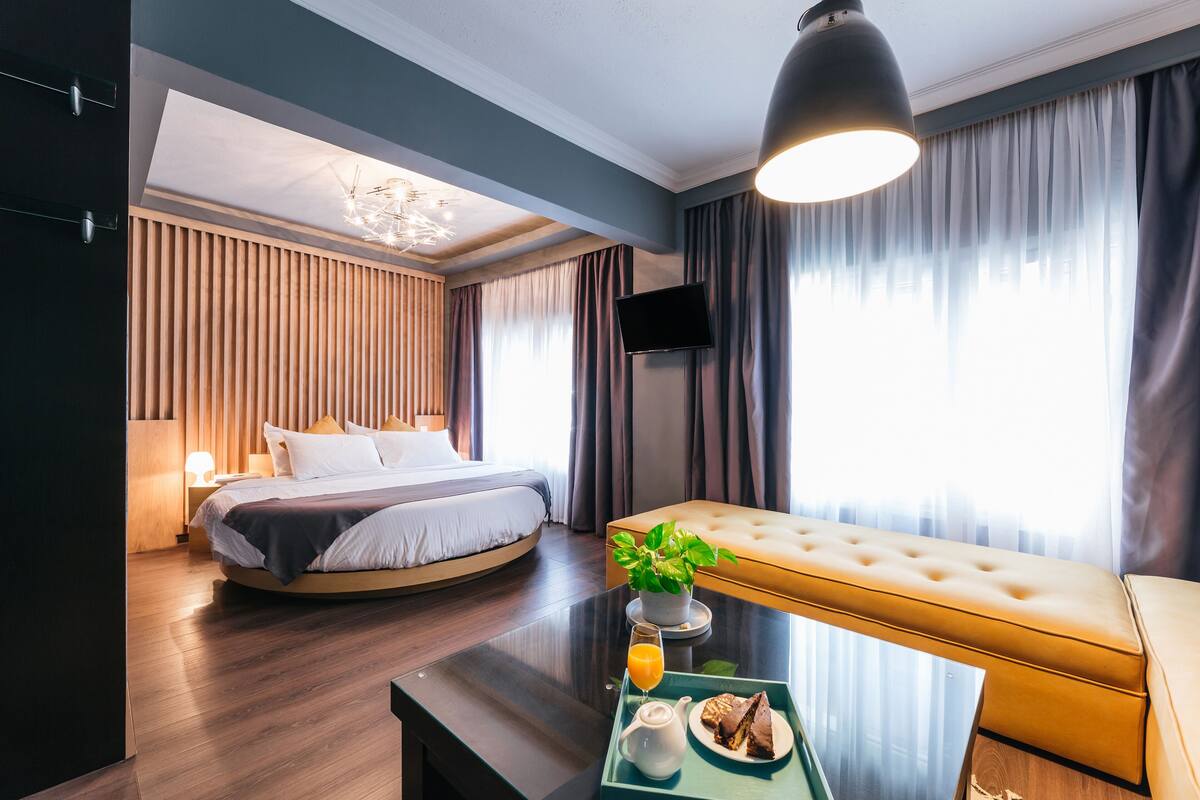 A modern hotel room with a bed, a sofa, a dining table with breakfast, and a TV.