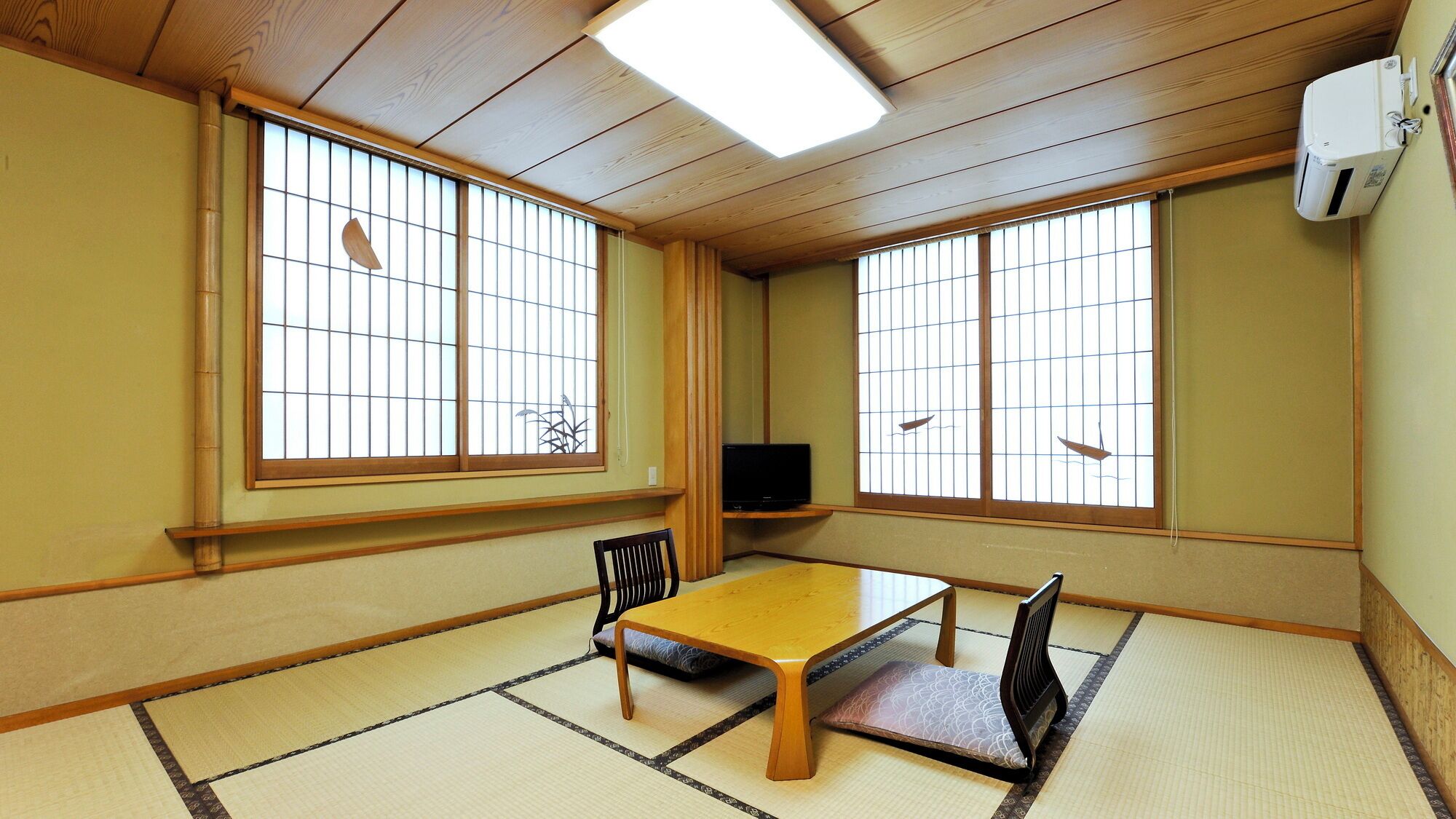 A traditional Japanese room with tatami flooring, a low wooden table, two chairs, a television, and large windows with a view of the sky.