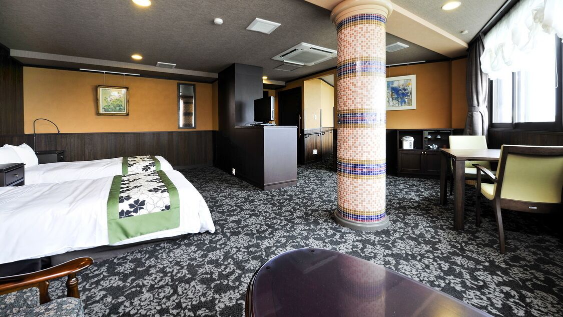A hotel room with a bed, a desk, a chair, a minibar, and a large pillar.