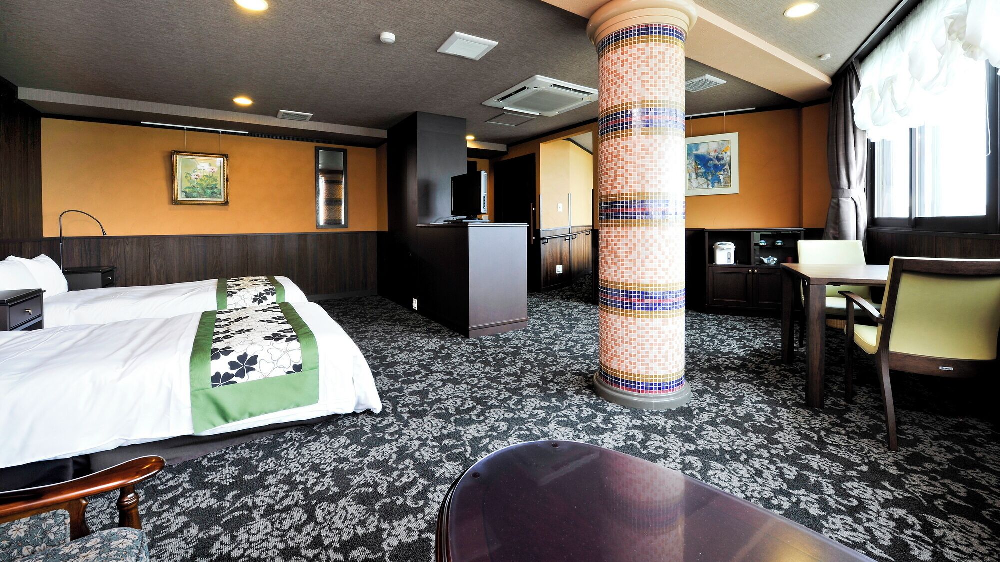 A hotel room with a bed, a desk, a chair, a pillar, and a kitchenette.